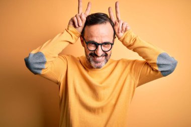 Orta yaşlı, ağarmış, günlük süveter ve gözlük takan, izole edilmiş sarı arka planda tuhaf ve çılgın pozlar veren, parmakları kafasında tavşan kulağı olan, neşeli gülümseyen bir adam.