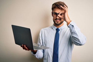 Young businessman wearing glasses working using laptop standing over white background with happy face smiling doing ok sign with hand on eye looking through fingers