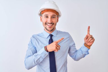 Young business man wearing contractor safety helmet over isolated background smiling and looking at the camera pointing with two hands and fingers to the side.