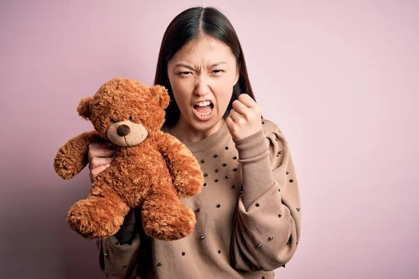 Young asian woman hugging teddy bear stuffed animal over pink isolated background annoyed and frustrated shouting with anger, crazy and yelling with raised hand, anger concept