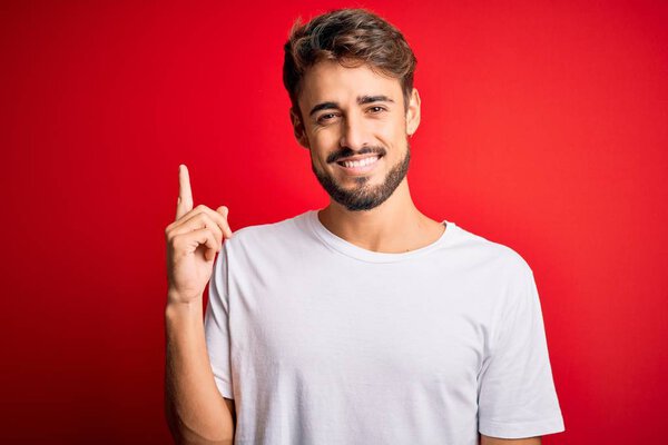 Young handsome man with beard wearing casual t-shirt standing over red background with a big smile on face, pointing with hand and finger to the side looking at the camera.