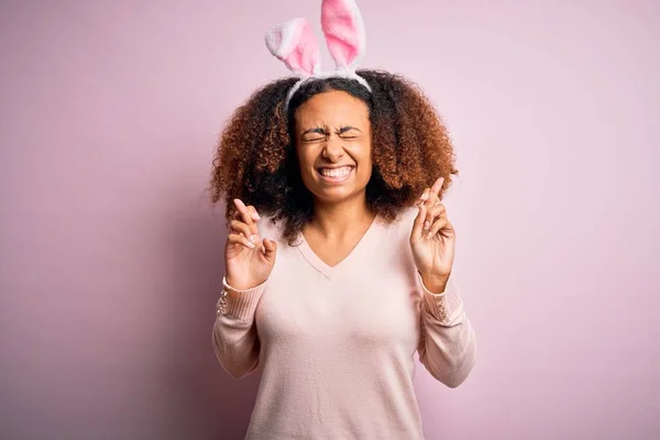 Young african american woman with afro hair wearing bunny ears over pink background gesturing finger crossed smiling with hope and eyes closed. Luck and superstitious concept.