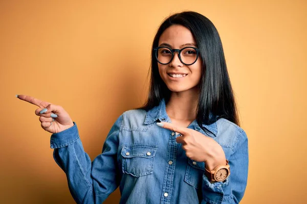 Young beautiful chinese woman wearing casual denim shirt over isolated yellow background smiling and looking at the camera pointing with two hands and fingers to the side.