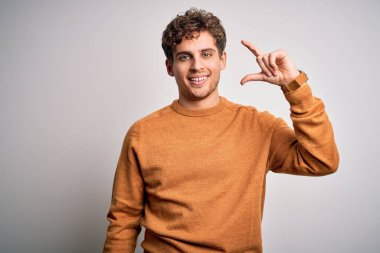 Young blond handsome man with curly hair wearing casual sweater over white background smiling and confident gesturing with hand doing small size sign with fingers looking and the camera. Measure concept.