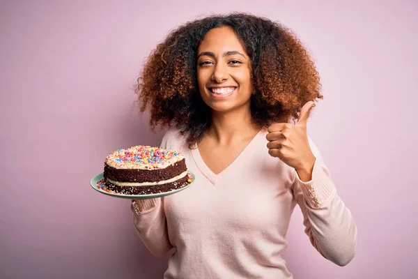Young african american woman with afro hair holding delicious birthday cake happy with big smile doing ok sign, thumb up with fingers, excellent sign