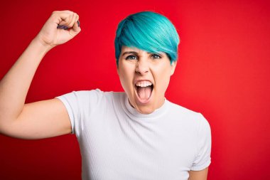 Young beautiful woman with blue fashion hair wearing casual t-shirt over red background angry and mad raising fist frustrated and furious while shouting with anger. Rage and aggressive concept.