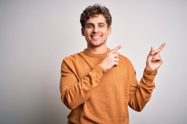 Young blond handsome man with curly hair wearing casual sweater over white background smiling and looking at the camera pointing with two hands and fingers to the side.