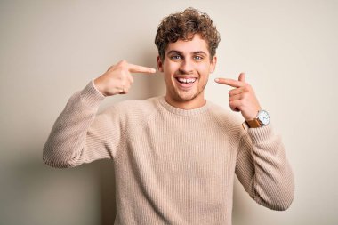 Young blond handsome man with curly hair wearing casual sweater over white background smiling cheerful showing and pointing with fingers teeth and mouth. Dental health concept.