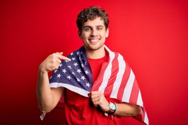 Young blond patriotic man with curly hair wearing USA flag celebrating independence day with surprise face pointing finger to himself