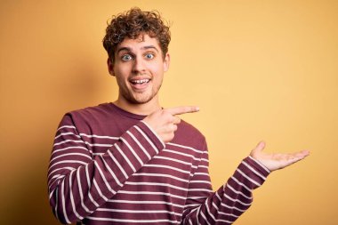 Young blond handsome man with curly hair wearing casual striped sweater amazed and smiling to the camera while presenting with hand and pointing with finger.