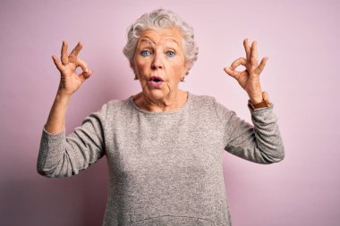Senior beautiful woman wearing casual t-shirt standing over isolated pink background looking surprised and shocked doing ok approval symbol with fingers. Crazy expression