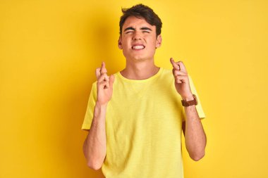 İzole edilmiş arka planda sarı t-shirt giyen genç çocuk umut ve gözler kapalı bir şekilde gülümseyerek işaret ediyor. Şans ve batıl inanç..