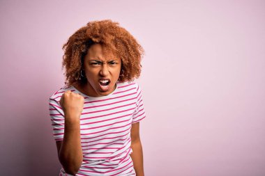 Young beautiful African American afro woman with curly hair wearing casual striped t-shirt angry and mad raising fist frustrated and furious while shouting with anger. Rage and aggressive concept.
