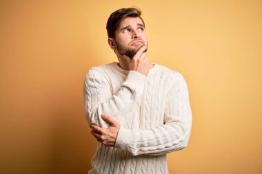 Young blond man with beard and blue eyes wearing white sweater over yellow background with hand on chin thinking about question, pensive expression. Smiling with thoughtful face. Doubt concept.