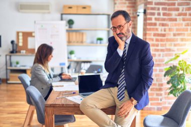 Middle age handsome businessman wearing glasses sitting on desk at the office thinking looking tired and bored with depression problems with crossed arms.
