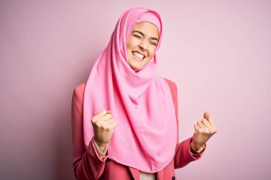 İzole edilmiş pembe arka planda duran Müslüman tesettürlü genç ve güzel bir kız çok mutlu ve heyecanlı bir şekilde kollarını kaldırıp, gülümseyerek ve başarı için çığlık atarak zafer jesti yapıyor. Kutlama konsepti.