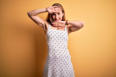 Young beautiful blonde woman on vacation wearing summer dress over yellow background Smiling cheerful playing peek a boo with hands showing face. Surprised and exited