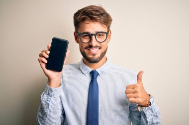 Young businessman holding smartphone standing over isolated white background happy with big smile doing ok sign, thumb up with fingers, excellent sign