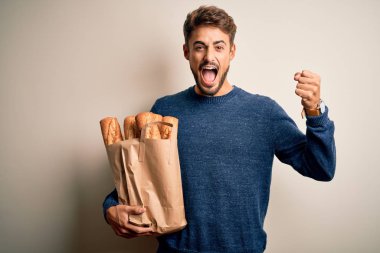 Young man holding paper bag with bread standing over isolated red bakground screaming proud and celebrating victory and success very excited, cheering emotion