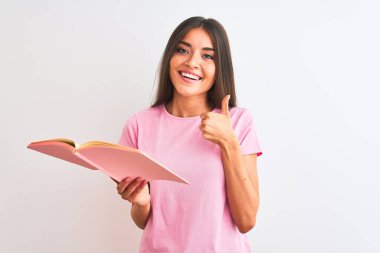 Young beautiful student woman reading book standing over isolated white background happy with big smile doing ok sign, thumb up with fingers, excellent sign