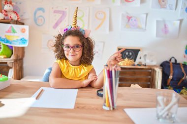 Beautiful toddler wearing glasses and unicorn diadem sitting on desk at kindergarten with a big smile on face, pointing with hand and finger to the side looking at the camera.