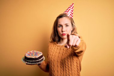 Young beautiful blonde woman holding birthday cake over isolated yellow background pointing with finger to the camera and to you, hand sign, positive and confident gesture from the front