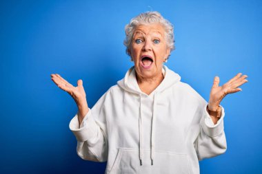 Senior beautiful sporty woman wearing white sweatshirt over isolated blue background celebrating crazy and amazed for success with arms raised and open eyes screaming excited. Winner concept