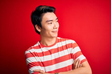 Young handsome chinese man wearing casual striped t-shirt standing over red background looking to the side with arms crossed convinced and confident