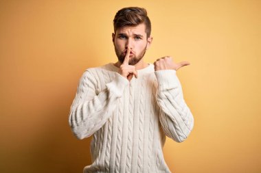 Young blond man with beard and blue eyes wearing white sweater over yellow background asking to be quiet with finger on lips pointing with hand to the side. Silence and secret concept.