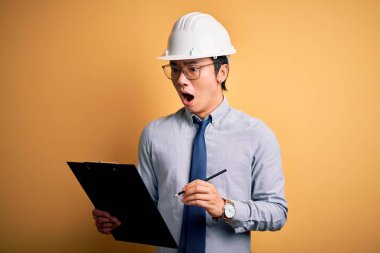 Young handsome chinese engineer man wearing safety helmet holding clipboard scared in shock with a surprise face, afraid and excited with fear expression