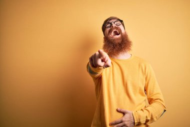 Sarıdan izole edilmiş arka planda gözlüklü, yakışıklı, kızıl saçlı ve sakallı bir adam. Sana gülüyor, eli vücudunun üzerinde kamerayı işaret ediyor. Utanç ifadesi.