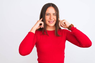 Young beautiful girl wearing red casual t-shirt standing over isolated white background covering ears with fingers with annoyed expression for the noise of loud music. Deaf concept.