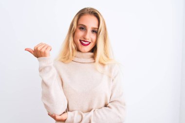 Balıkçı yaka kazak giyen genç ve güzel bir kadın izole edilmiş beyaz arka planda gülümsüyor ve başparmağıyla yan tarafa bakıyor..