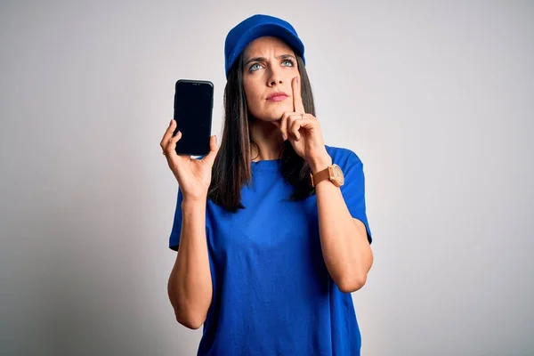 Young delivery woman with blue eyes wearing cap holding smartphone serious face thinking about question, very confused idea