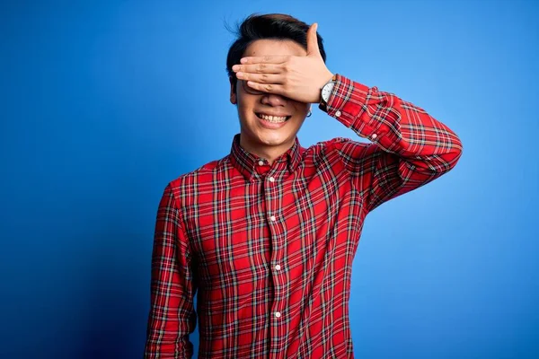 Young handsome chinese man wearing casual shirt and glasses over blue background smiling and laughing with hand on face covering eyes for surprise. Blind concept.