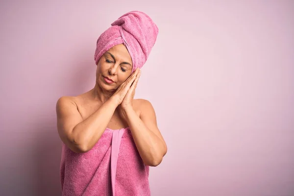 Middle age woman wearing bath towel from beauty body care over pink background sleeping tired dreaming and posing with hands together while smiling with closed eyes.