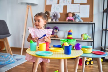 Anaokulunun masasında çatal bıçak ve yemek oyuncaklarıyla oynayan genç ve güzel bir çocuk.