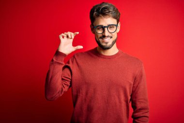 Sakallı, gözlüklü, süveter giyen, kırmızı arka planda duran, gülümseyen, kendine güvenen, el hareketi yapan, parmaklarıyla işaret eden ve kameraya bakan genç yakışıklı bir adam. Ölçü kavramı.