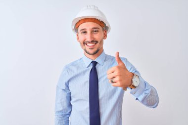 Young business man wearing contractor safety helmet over isolated background doing happy thumbs up gesture with hand. Approving expression looking at the camera showing success.