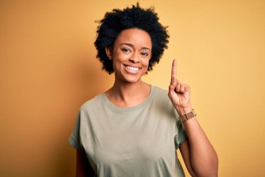 Young beautiful African American afro woman with curly hair wearing casual t-shirt showing and pointing up with finger number one while smiling confident and happy.
