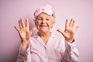 İzole edilmiş pembe arka planda uyku maskesi ve pijama giyen son sınıf güzeli bir kadın on numarayı işaret ederken kendinden emin ve mutlu bir şekilde gülümsüyor..