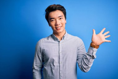 Genç, yakışıklı, sıradan bir gömlek giyen, mavi arka plan üzerinde duran, beş numaralı parmaklarıyla işaret eden, kendinden emin ve mutlu gülümseyen Çinli bir adam..