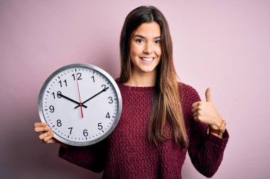 Geri sayım yapan güzel bir kız pembe arka planda büyük bir saat tutuyor. Gülümsüyor ve gülümsüyor. İşaret veriyor.