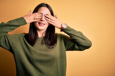 Young brunette woman with blue eyes wearing green casual sweater over yellow background covering eyes with hands smiling cheerful and funny. Blind concept.