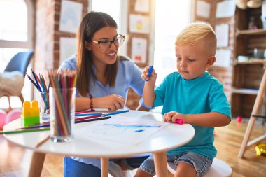 Genç beyaz çocuk öğretmeniyle oyun okulunda oynuyor. Anne ve oğlu oyun odasında renkli kalemlerle beraberlik çiziyor.