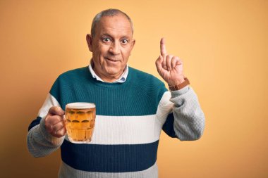 Üst düzey yakışıklı adam izole edilmiş sarı arka planda duran bira kavanozunu içerken yüzünde kocaman bir gülümsemeyle kameraya el ile işaret ediyor..