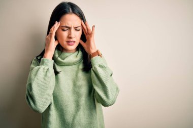 Young brunette woman with blue eyes wearing turtleneck sweater over white background with hand on head for pain in head because stress. Suffering migraine.