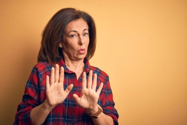 Middle age beautiful woman wearing casual shirt standing over isolated yellow background Moving away hands palms showing refusal and denial with afraid and disgusting expression. Stop and forbidden.