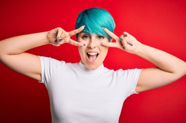 Mavi saçlı, kırmızı arka planda sıradan tişörtler giyen genç ve güzel bir kadın. Barış sembolü yapıyor. Yüzünde parmaklar, gülümseyerek zaferi gösteriyor.