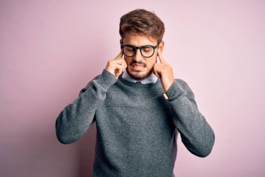 Sakallı, gözlüklü ve süveterli genç bir adam pembe arka planda duruyor. Yüksek sesli müzik sesi için kulaklarını parmaklarıyla kapatıyor. Sağır kavramı.